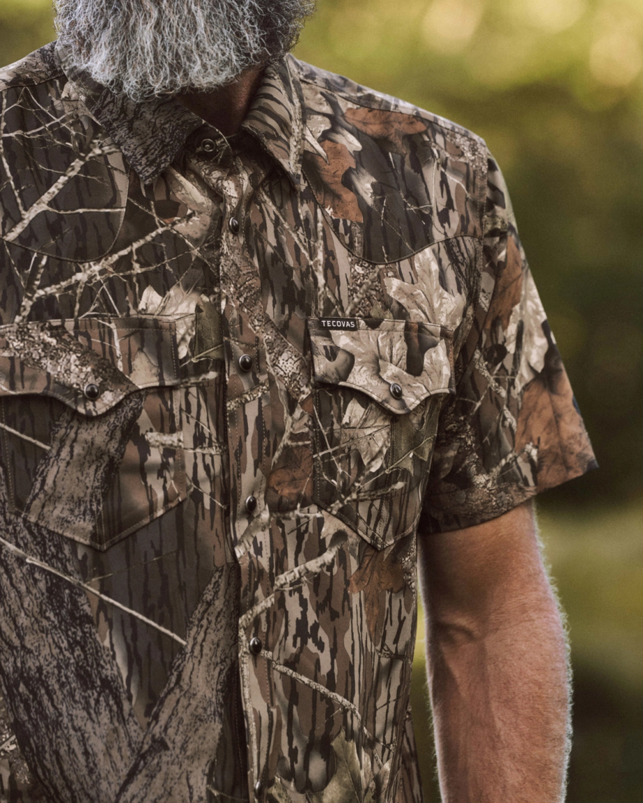 A person with a gray beard wears a short-sleeve, camouflage-pattern button-up shirt with double chest pockets outdoors.