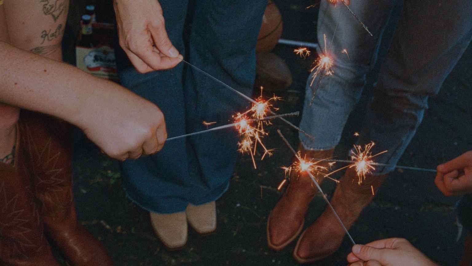 Four people wearing Tecovas boots stand close together holding lit sparklers, with their hands extended toward the center of the group.