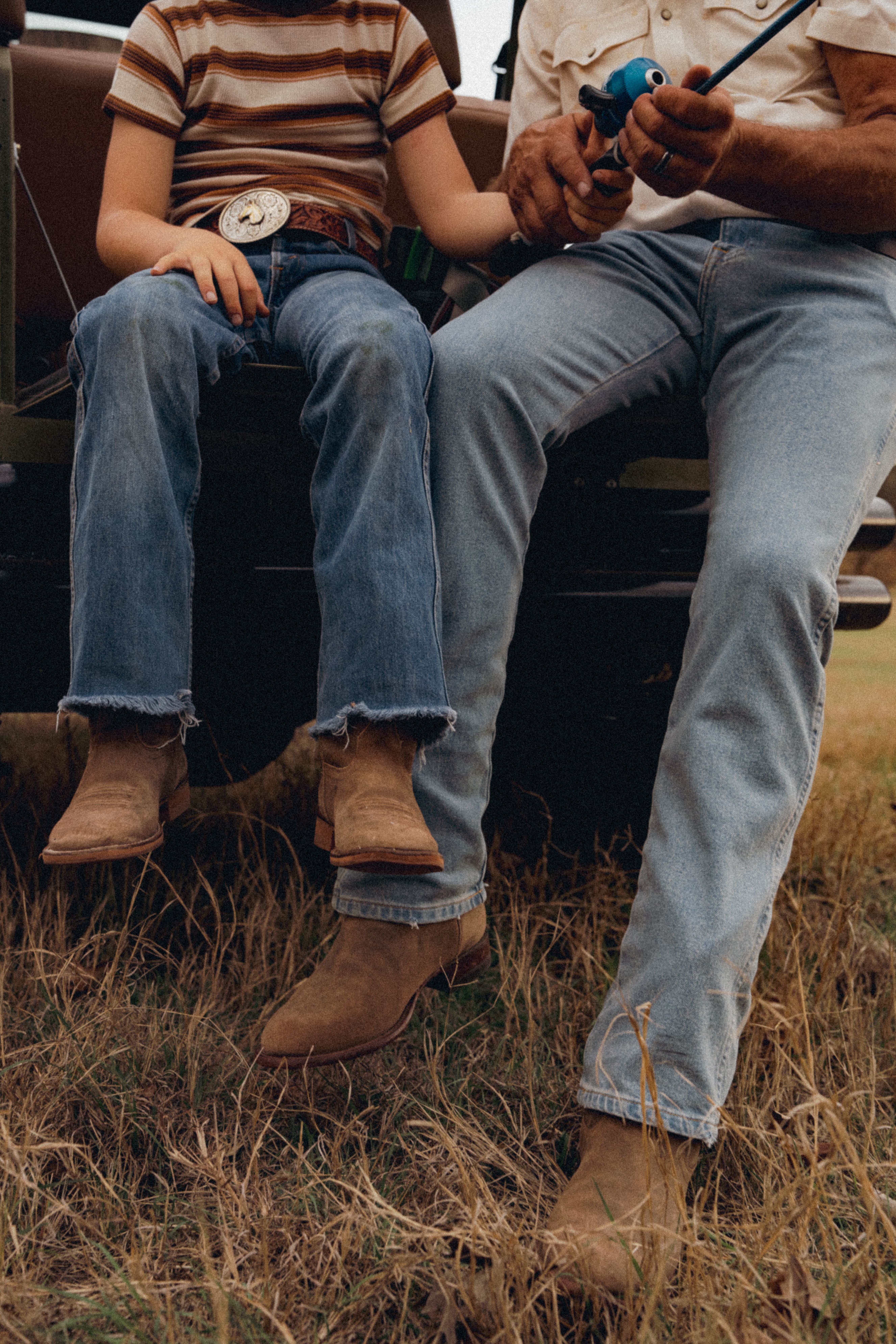 A child and an adult sit on the tailgate of a vehicle in a field, holding a fishing rod. Both wear jeans, boots, and casual shirts. The child has a large belt buckle.