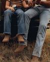 A child and an adult sit on the tailgate of a vehicle in a field, holding a fishing rod. Both wear jeans, boots, and casual shirts. The child has a large belt buckle.