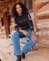 A woman with curly hair wearing a black sweater and blue jeans sits on a porch bench outside a rustic wooden cabin. Red chili peppers hang from the wall behind her.