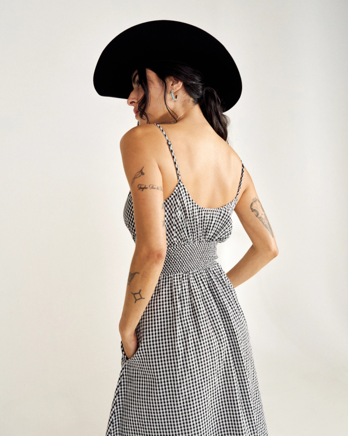 A woman in a black hat wearing a sleeveless checkered dress, showing her back and tattoos on her arms, stands against a plain background.