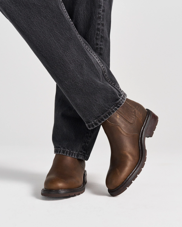 The Chelsea in Cafe Cowhide. Person wearing dark denim jeans and brown leather boots from The Chelsea Cowhide stands on a white background with one leg crossed in front of the other.