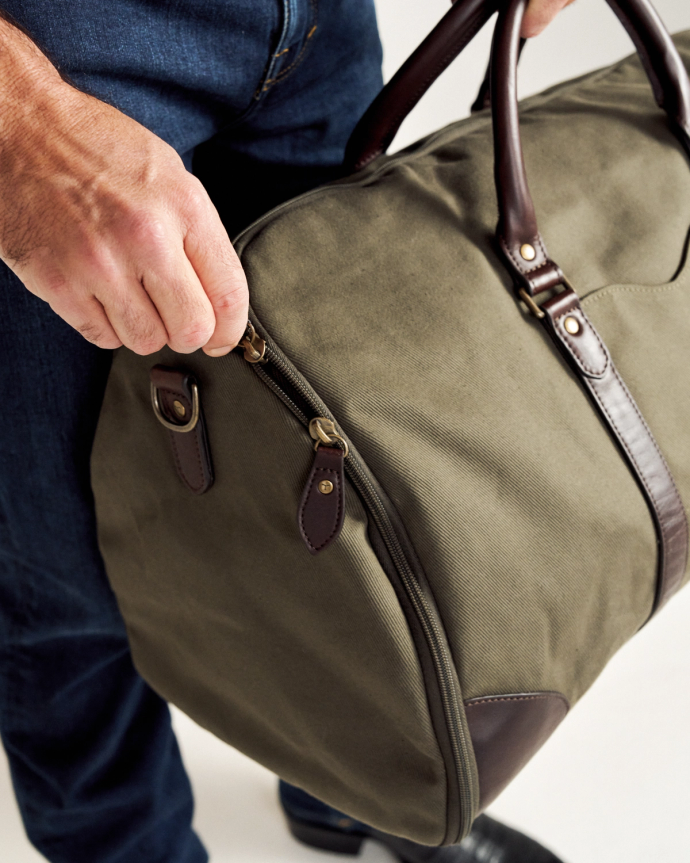 Person holding a green canvas duffel bag with brown leather straps and zipper details.