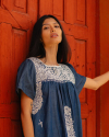 A woman with long dark hair, wearing a blue and white embroidered dress, stands against a red wooden door.