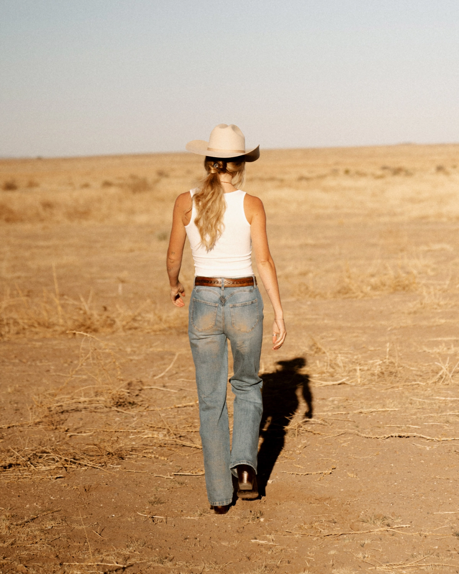 A person wearing a cowboy hat, white tank top, and blue jeans walks across a dry, open field with sparse vegetation.