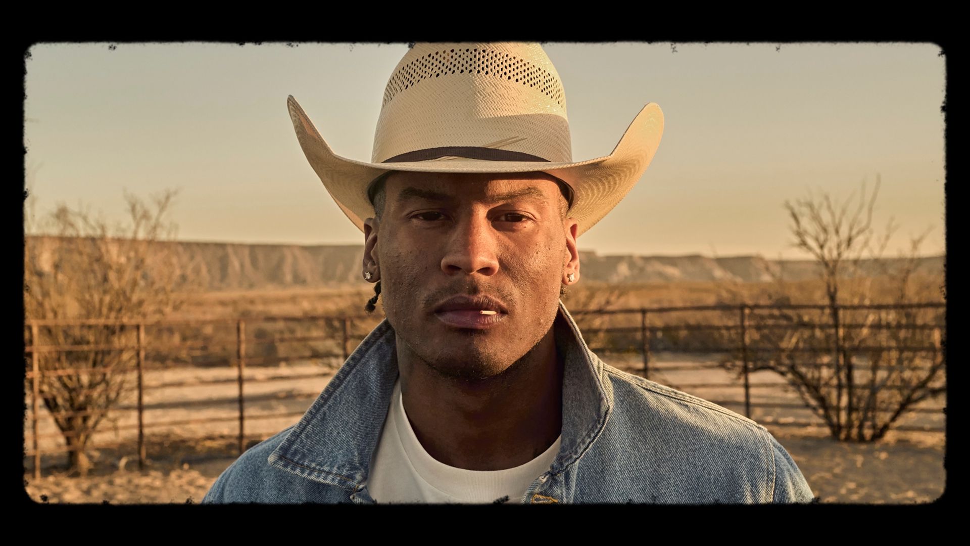 A man wearing a white cowboy hat and denim jacket stands outdoors in a desert landscape with dry bushes and distant hills.