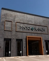 Exterior view of the Pinto Ranch store with a brick facade, large sign, and orange-framed entrance under a clear blue sky.