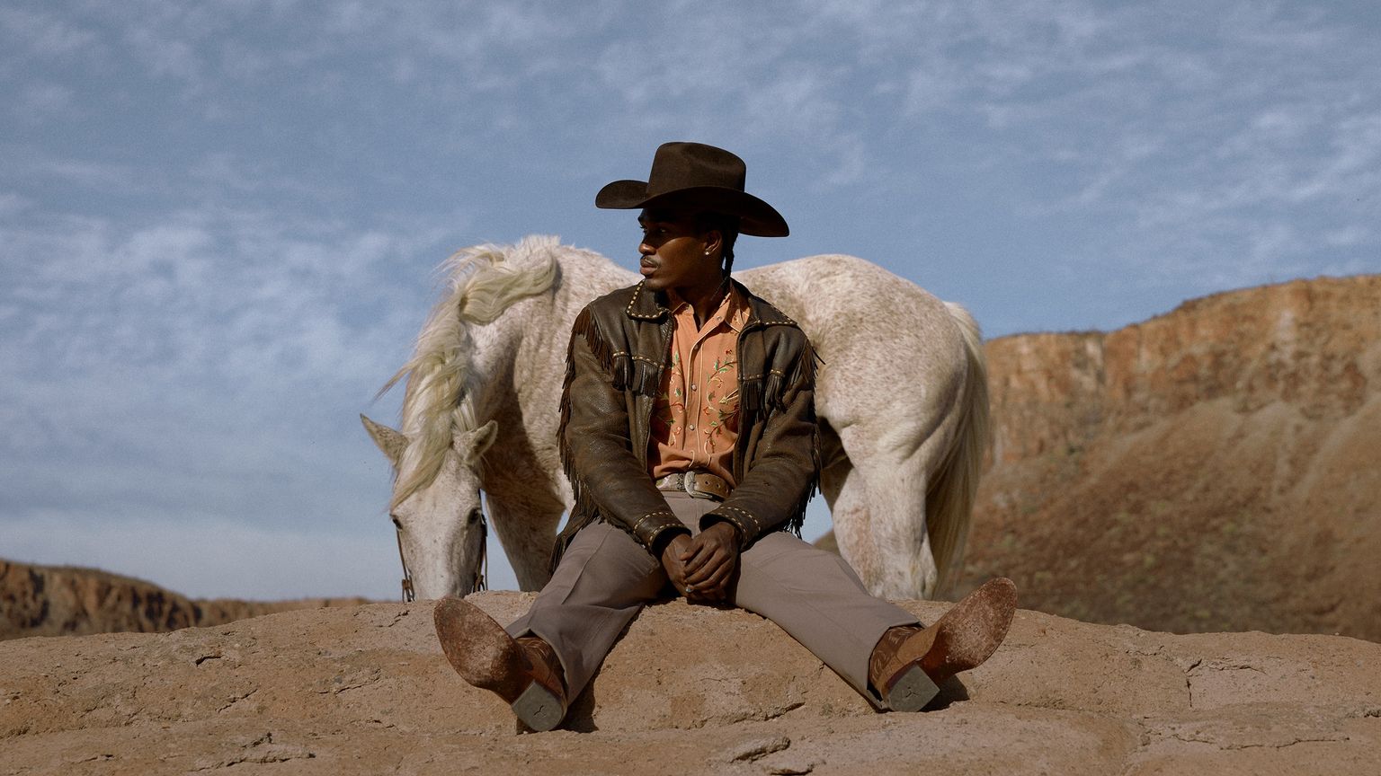 A person wearing a cowboy hat and jacket sits on a rock, with a white horse standing behind them against a backdrop of sky and cliffs.