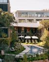 Outdoor pool area with loungers and umbrellas surrounded by greenery. Modern building in the background with large windows and multiple stories.