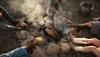 People wearing cowboy boots sit around a campfire on rocky ground, smoke rising from the flames.
