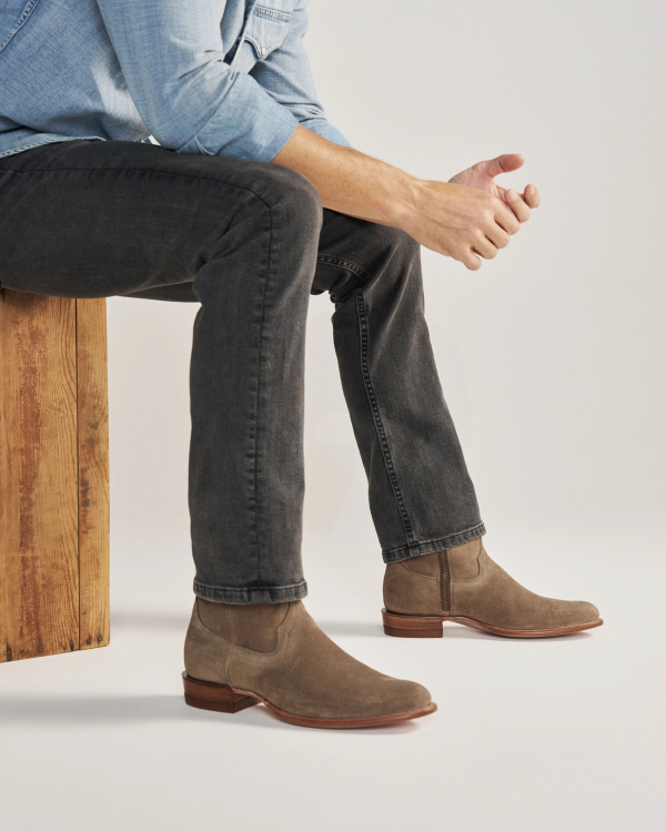 The Dean in Granite Suede. A man sitting on a wooden bench wears a light blue shirt, black jeans, and The Dean Suede Chelsea boots in taupe.