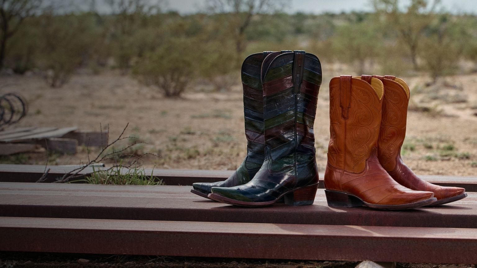 Two pairs of cowboy boots, one brown and one multicolored, are placed side by side on metal rails in an outdoor, desert-like setting.