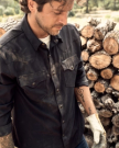 A man in a dirty work shirt and gloves stands next to a stack of cut logs, with tattoos visible on his forearm.