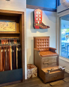 A well-organized shop corner featuring a variety of leather belts on display, a painting above, and a boot shining station with a cushioned seat and retro sign.