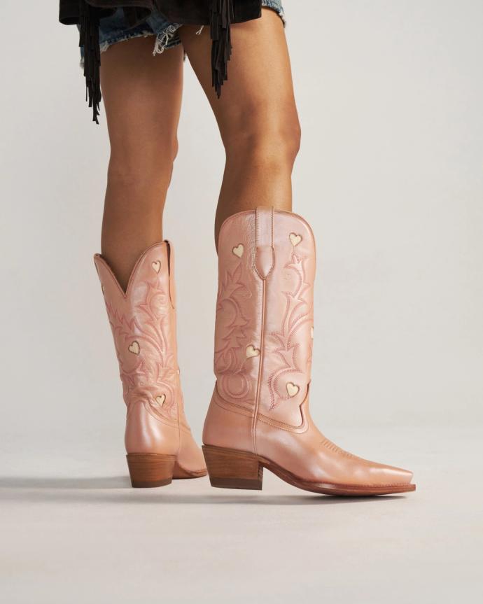 Person wearing light pink cowboy boots with heart-shaped cutouts, standing on a plain surface; only legs and lower outfit visible.