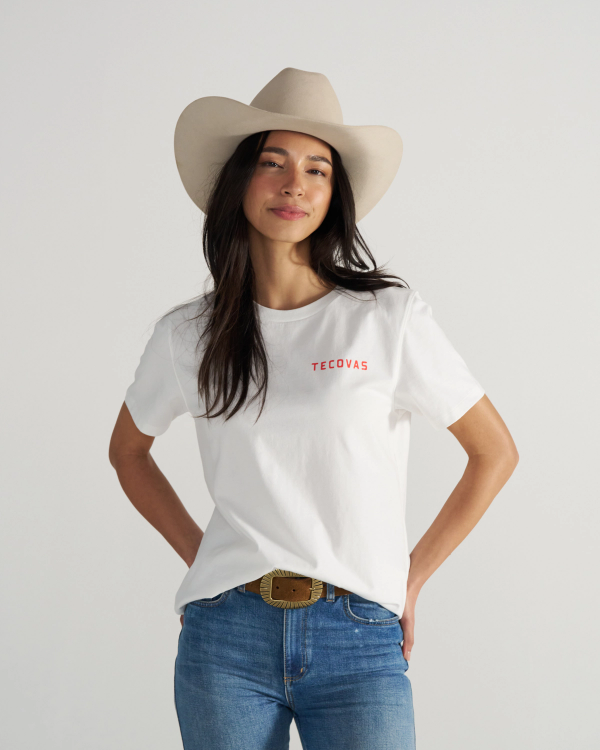 A woman in a beige cowboy hat, white "Tecovas" t-shirt, and blue jeans stands with hands in pockets against a plain light background.
