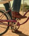 A person wearing jeans and brown boots stands with one foot on the pedal of a vintage purple bicycle labeled “Fair Lady” on a dirt surface.