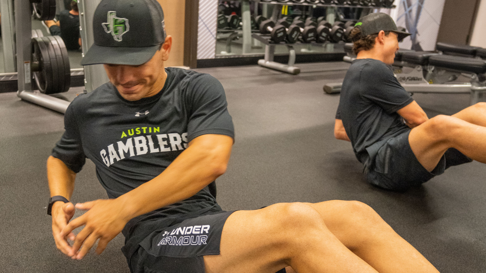 Two individuals are doing Russian twists on a gym floor; one is in focus, wearing Austin Gamblers apparel. Weight racks are visible in the background.