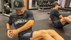 Two individuals are doing Russian twists on a gym floor; one is in focus, wearing Austin Gamblers apparel. Weight racks are visible in the background.