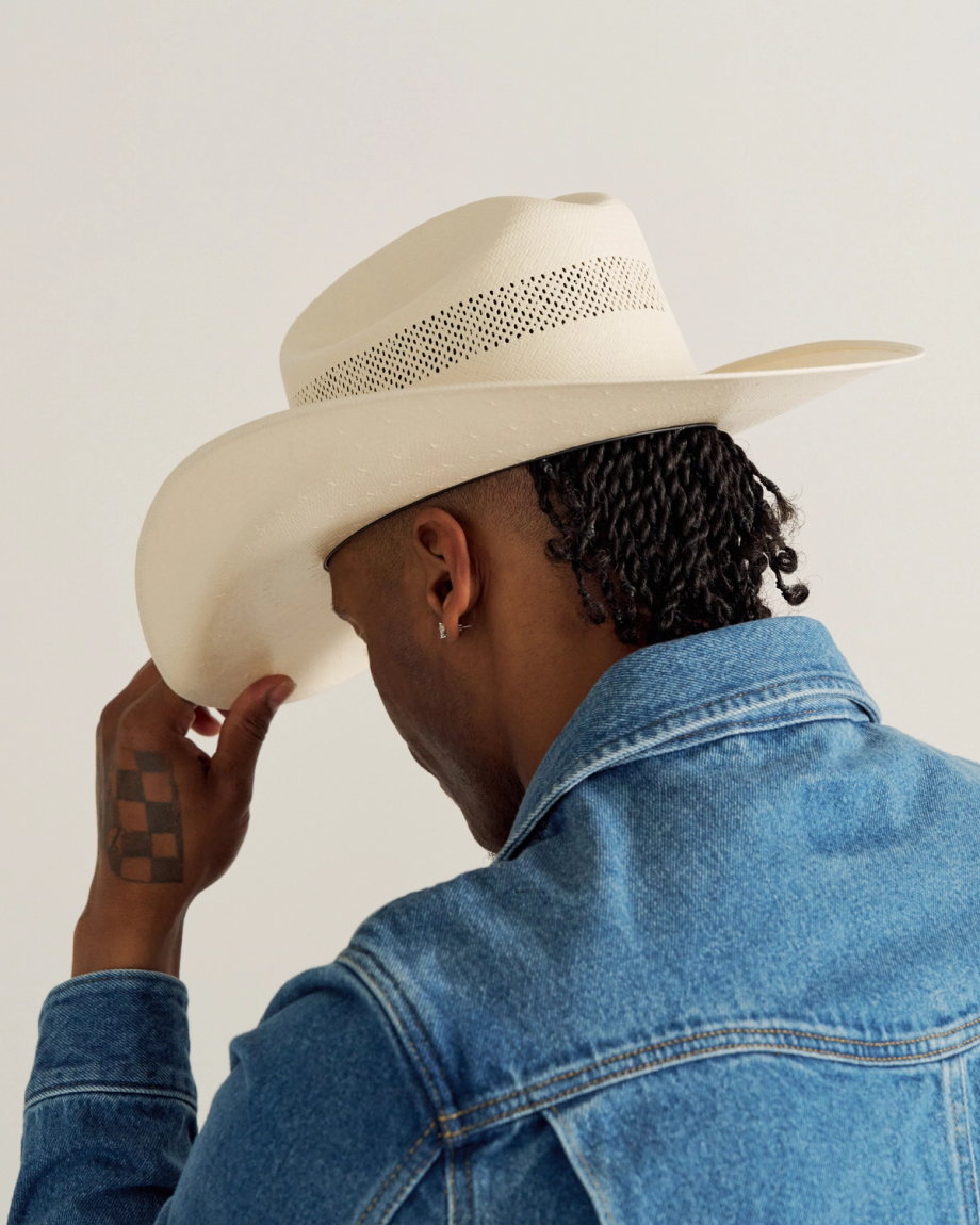 Person in a denim jacket and cream-colored cowboy hat, viewed from behind, holding the brim of the hat with one hand; checkered tattoo visible on wrist.