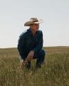 A man wearing a cowboy hat and denim clothing kneels in a grassy field under a clear sky.