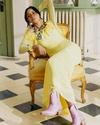 Woman in a long yellow dress and pink boots sits on a yellow armchair in a sunlit room with checkered floor tiles.