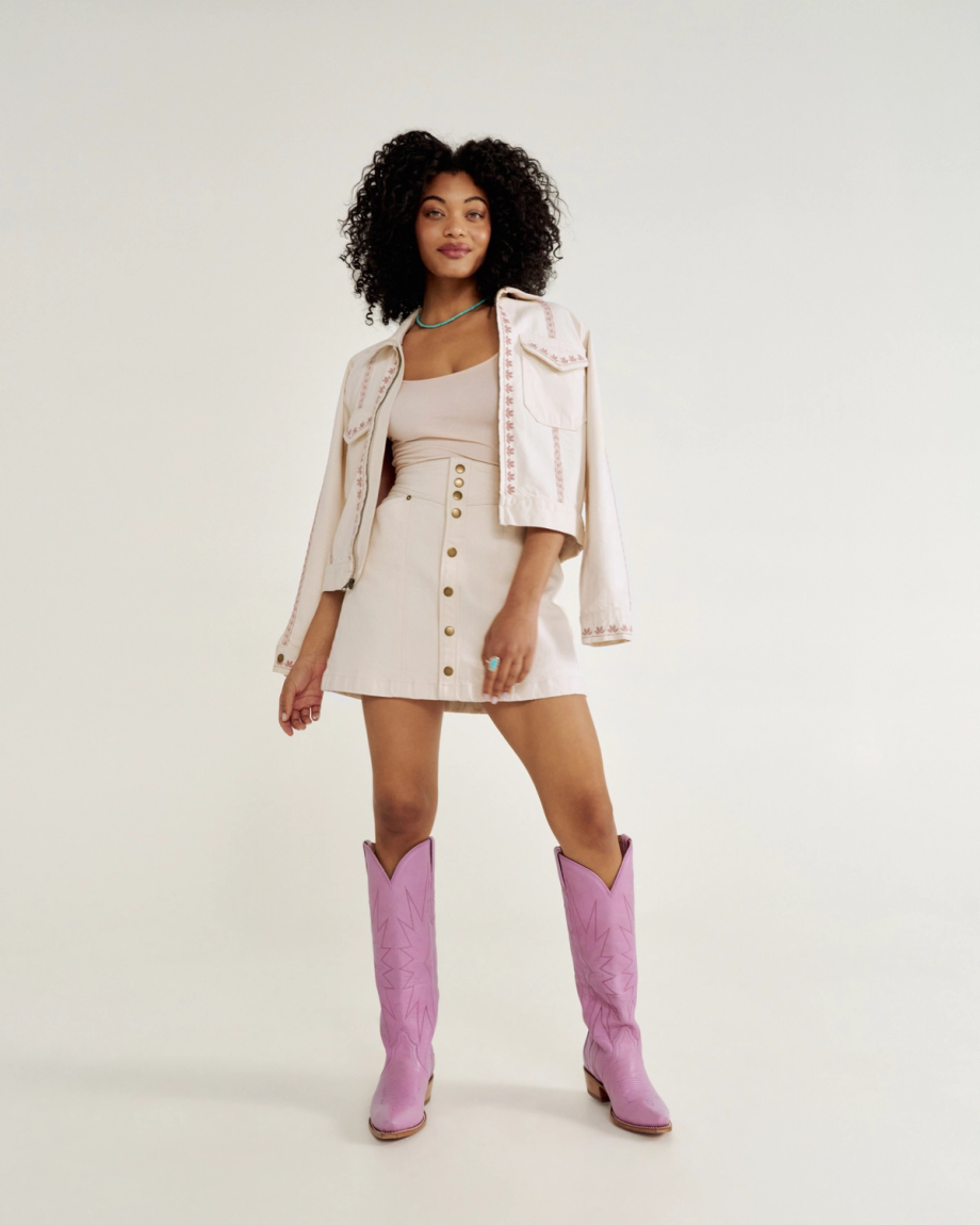 A person with curly hair wearing a light-colored skirt, jacket, and tank top, paired with pink cowboy boots, stands against a plain background.