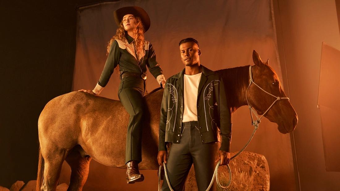 Two children in cowboy boots and jeans stand facing each other; one places a white cowboy hat on the other's head.