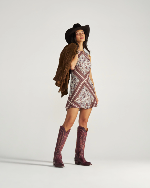 The Abby in Wine Goat. A woman in a cowboy hat, patterned dress, and boots stands holding a brown fringed jacket over her shoulder against a plain background, capturing the free-spirited style of The Abby Goat.