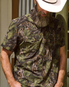 A person with a beard and hat wears a short-sleeve, green camouflage shirt, standing outdoors with hands in pockets.