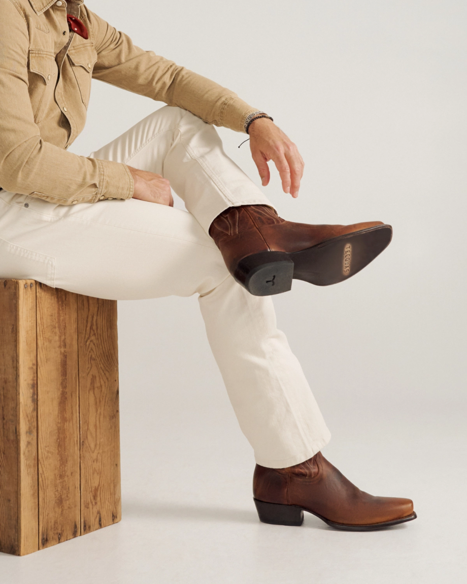 A person in a tan shirt and white pants sits on a wooden block with one leg crossed, wearing brown leather boots.