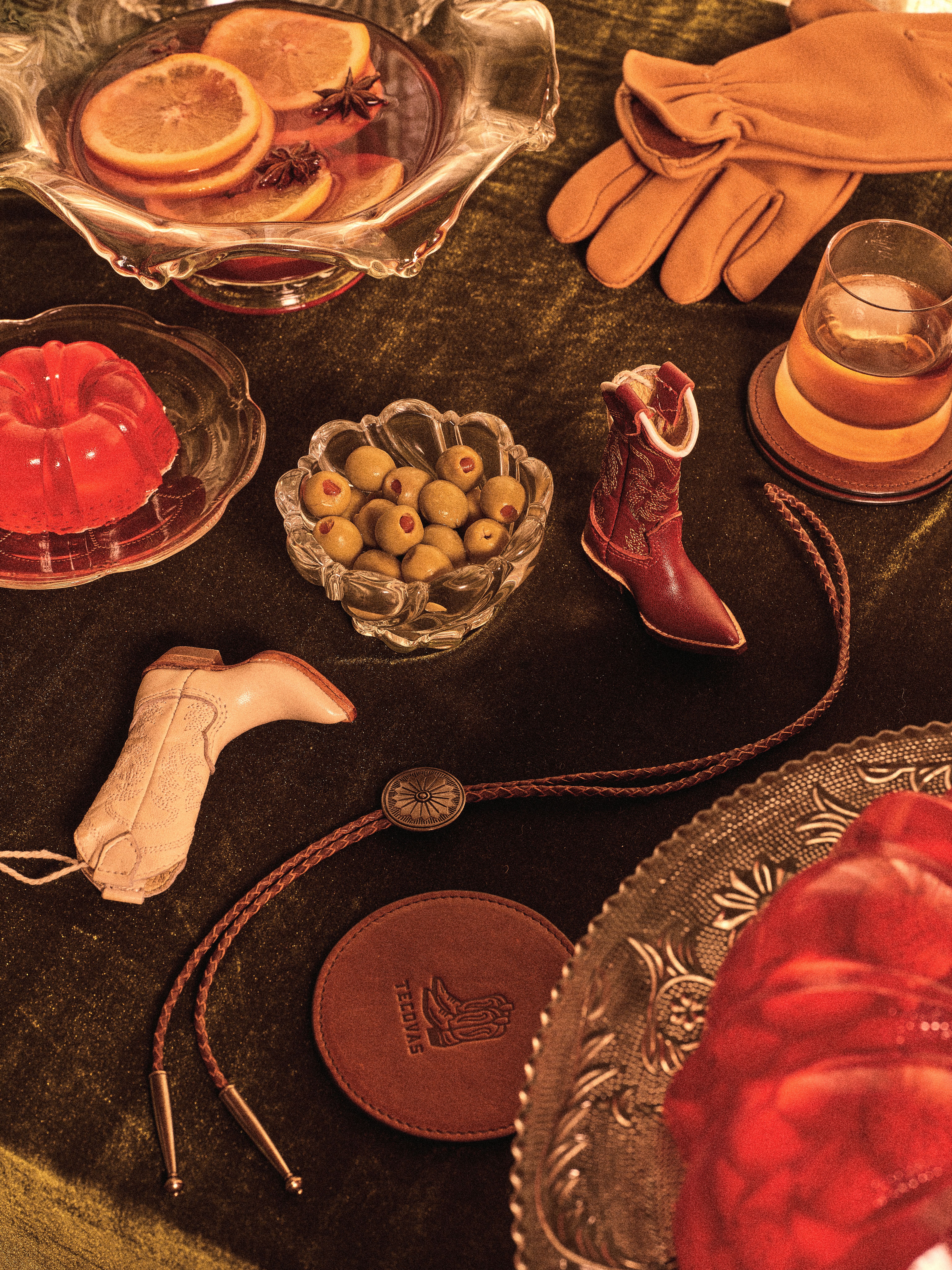 A table setting with gloves, olives, orange slices, a drink, two cowboy boot-shaped ornaments, a bolo tie, leather coaster, and red gelatin dessert on a velvet tablecloth.