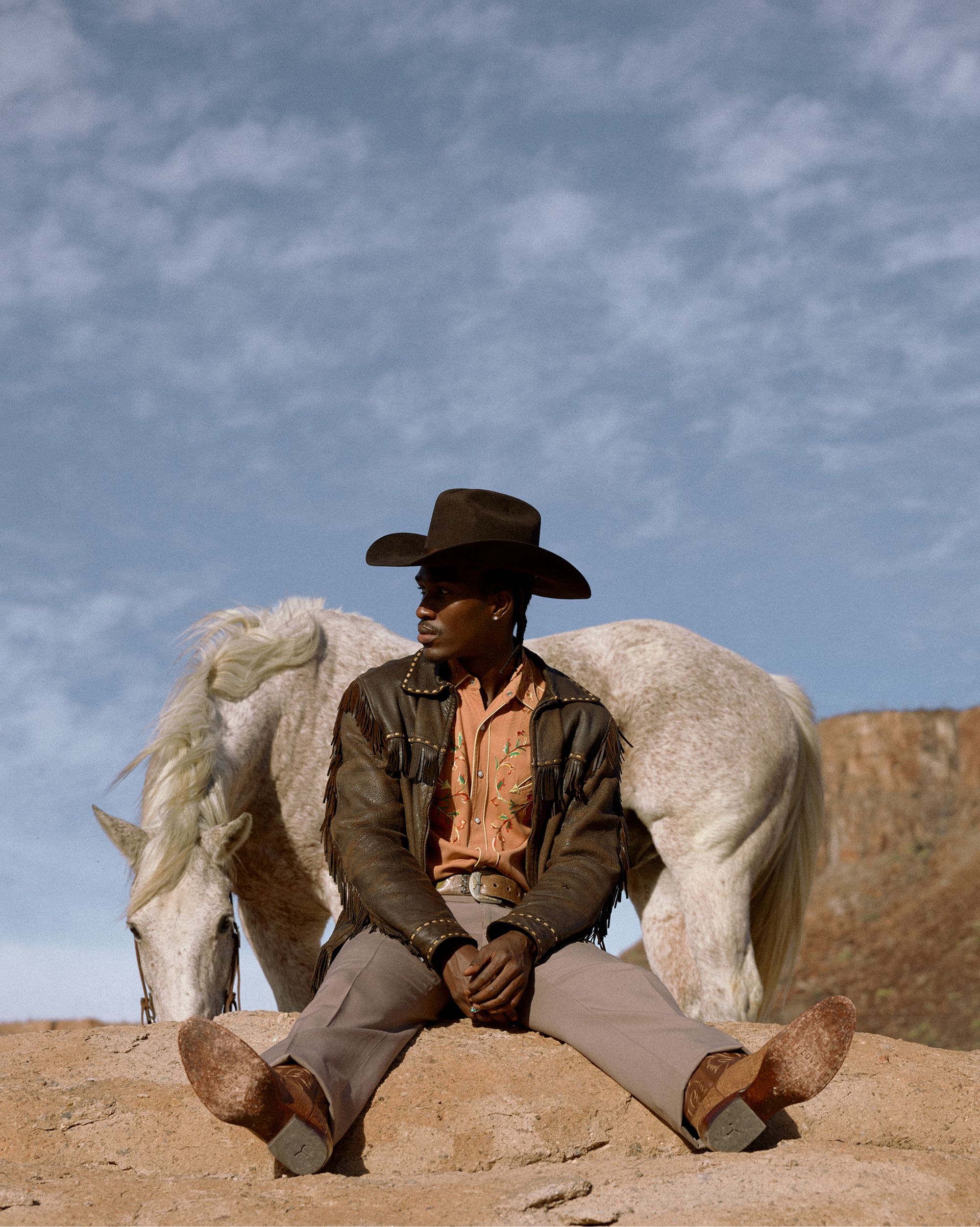 A person in a cowboy hat and fringe jacket sits on sand with legs outstretched; a white horse grazes behind them under a blue sky.