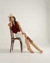 A woman wearing a cowboy hat, brown vest, white skirt, and tan cowboy boots sits sideways on a wooden chair against a plain background.