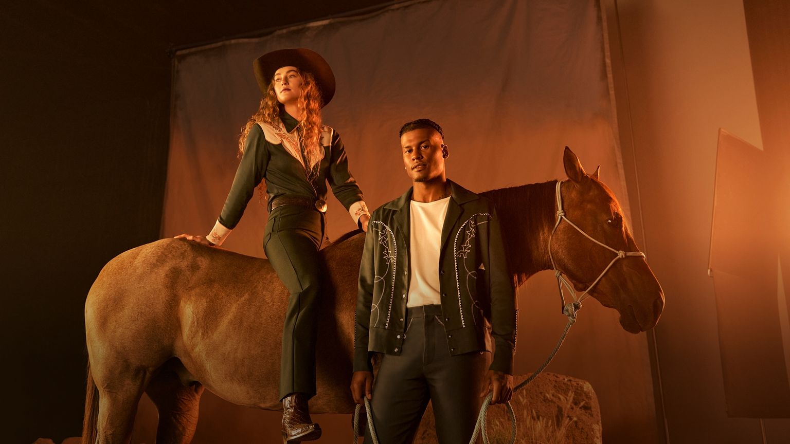 A woman in Western attire sits on a horse while a man in a black jacket stands beside them, holding the reins, with a plain backdrop and warm lighting.