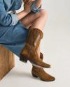 A person wearing a blue denim dress sits on a wooden bench, showing brown suede and leather cowboy boots with a low heel.