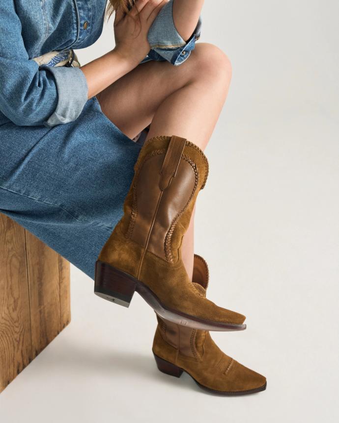 A person wearing a blue denim dress sits on a wooden bench, showing brown suede and leather cowboy boots with a low heel.
