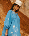 A woman in a blue dress with cactus and moon designs, wearing a white hat and large earrings, stands against a rocky background.