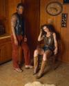 A man in a vest and corduroy pants stands next to a seated woman in a snakeskin top, shorts, and cowboy boots in a wood-paneled room with Western decor.