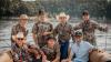 Seven men, some wearing cowboy hats and camouflage shirts, pose together on a boat with water and trees in the background.