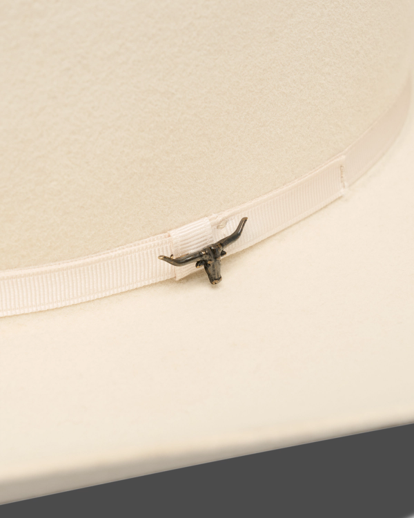 Close-up of a light-colored cowboy hat featuring a ribbon band with a small metallic longhorn emblem attached.