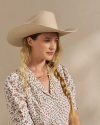 A person with long blond hair in a side braid wears a cream-colored cowboy hat and a floral-patterned blouse.