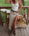 Person wearing a white tank top, long beige skirt with a high slit, red bandana, and brown boots stands on a wooden porch in front of a stack of firewood.