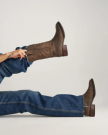 Person wearing blue jeans and brown cowboy boots lies outside The Tess Goat Cafe, one leg bent and raised, offering a stylish side view of the boots.