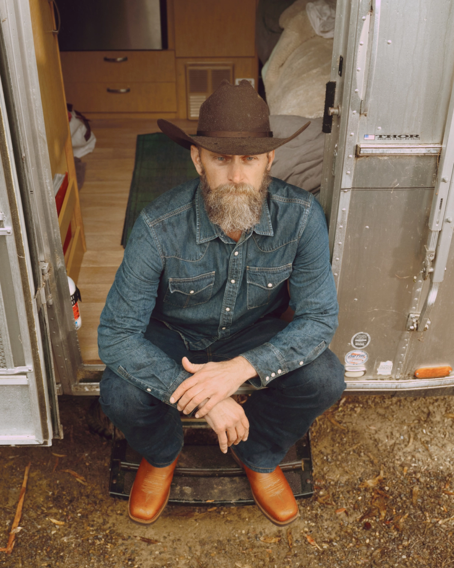 A person with a beard and a cowboy hat sits on steps at the entrance of a silver trailer. They wear a denim shirt, jeans, and brown boots, with the interior of the trailer visible behind them.
