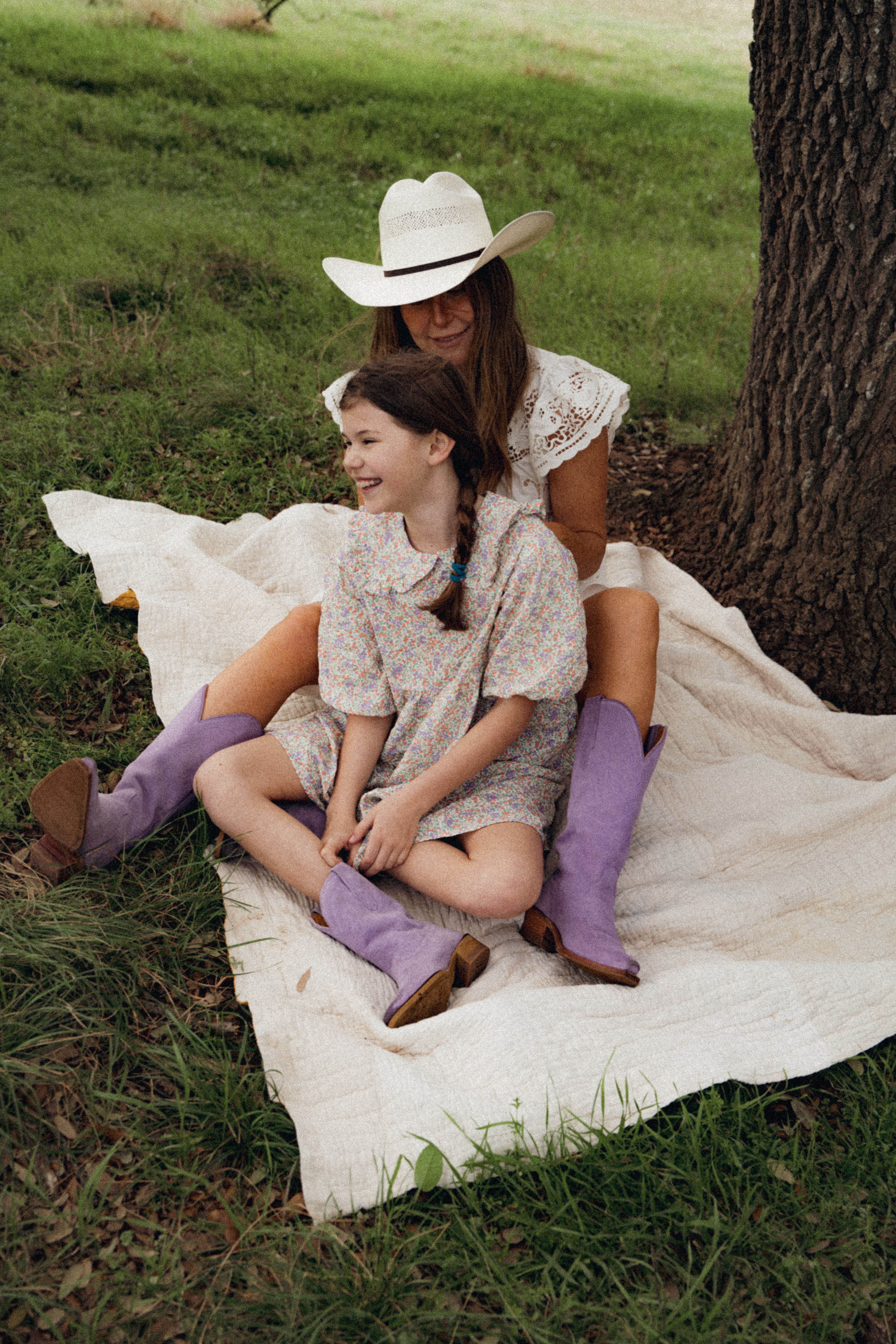 An adult and a child sit on a blanket under a tree, both wearing floral dresses and purple boots. The adult wears a white hat, and they are smiling.