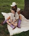 An adult and a child sit on a blanket under a tree, both wearing floral dresses and purple boots. The adult wears a white hat, and they are smiling.