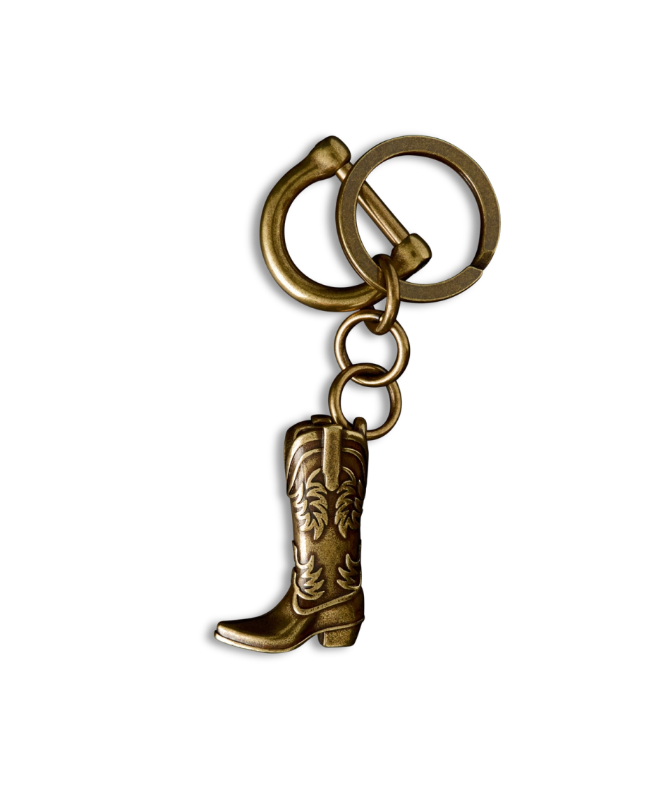 A brass keychain featuring a small, detailed cowboy boot charm with engraved patterns, set against a black background.