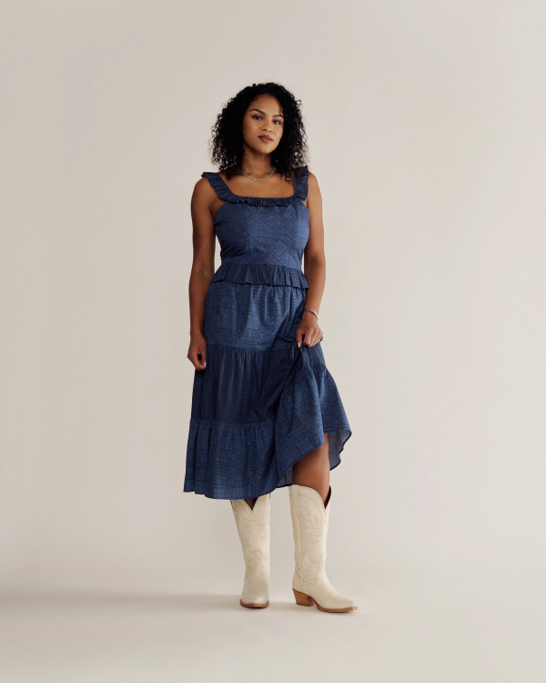 A woman in a blue sleeveless dress and white cowboy boots stands against a plain beige background, looking at the camera.